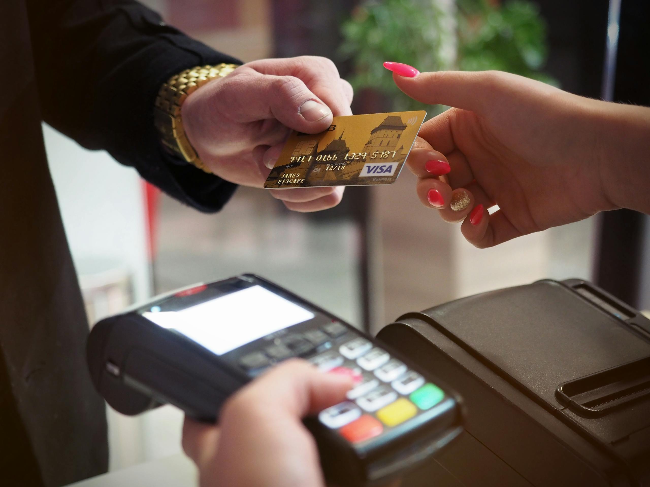 A white person's hand is holding a credit card on the right, which is passing the card to a white person's hand on the right with long red nails. The long nailed left hand is holding a credit card reader in the forefront.